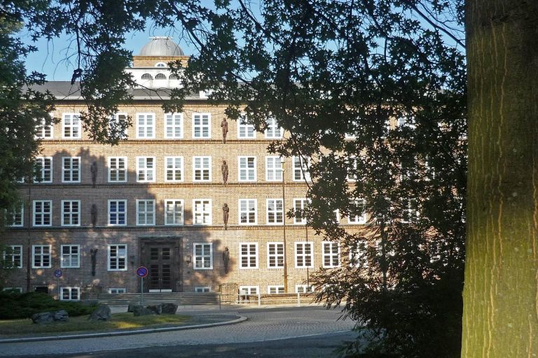 hinter einem großen Laubbaum sieht man einen mächtigen Klinkerbau Jünglingsfiguren an der Fassade